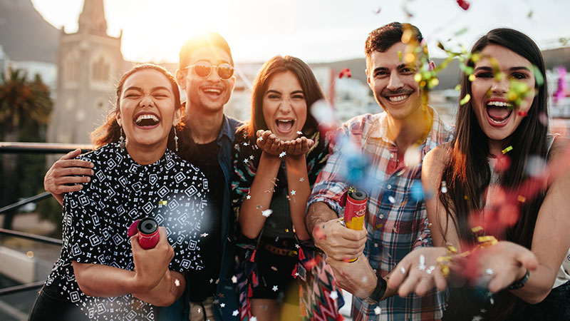 A group of Hispanic men and women laughing and throwing confetti as they celebrate outdoors.