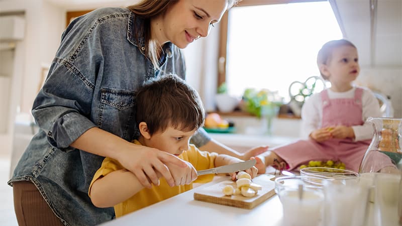 A smiling mom in a sun-lit kitchen helping her little boy cut slices of banana on a cutting board on her kitchen countertop, next to a little girl sitting on a high chair.