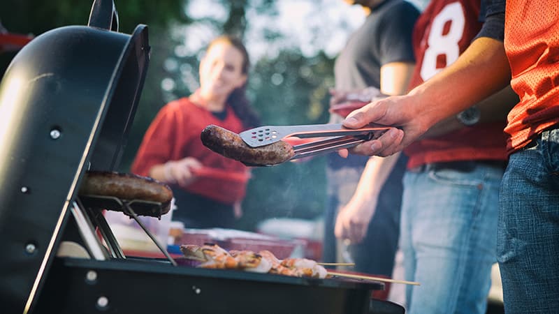 Four people cooking food at an outdoor grill, with one of them holding a grilled sausage with a pair of tongs.
