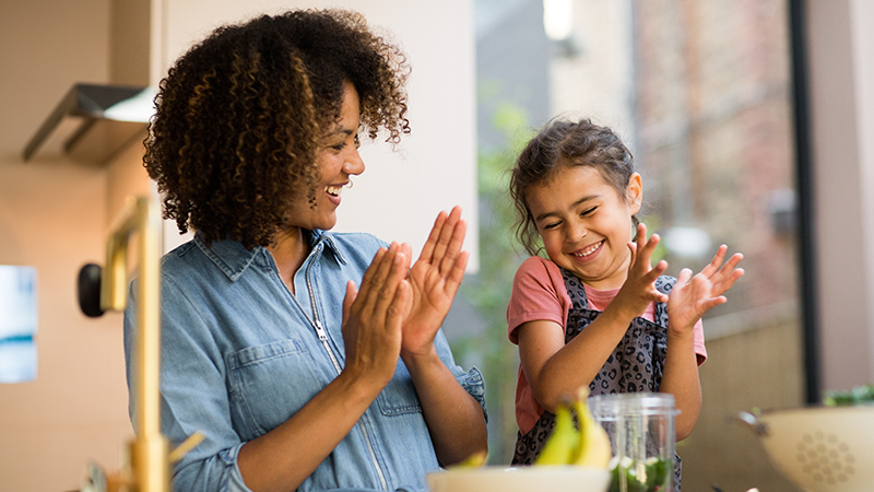 Smiling mom, dad and young daughter in a rustic kitchen in front of a counter with a salad bowl, a cutting board and a few scattered veggies and condiments.