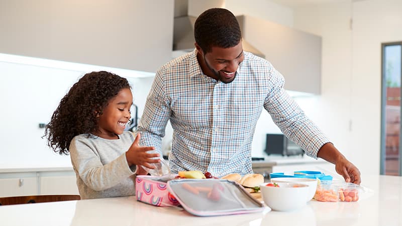 Father and young daughter standing at a modern kitchen counter, smiling with a pink lunch box, a white bowl of food and a small plastic bag of carrots in front of them.