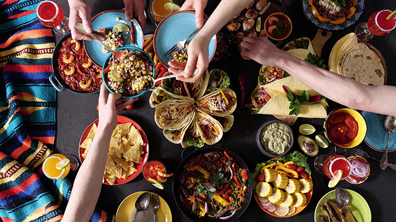 Four arms reaching over a collection of Mexican dishes, such as tacos, chips, tortillas, aguas frescas on a table over an indigenous themed tablecloth.