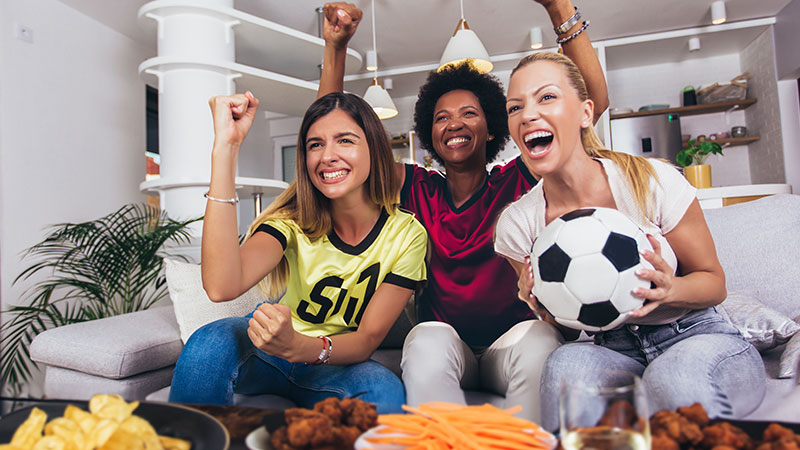 Three women wearing different team jerseys and cheering in front of a table full of game day snacks. One woman is holding a soccer ball.