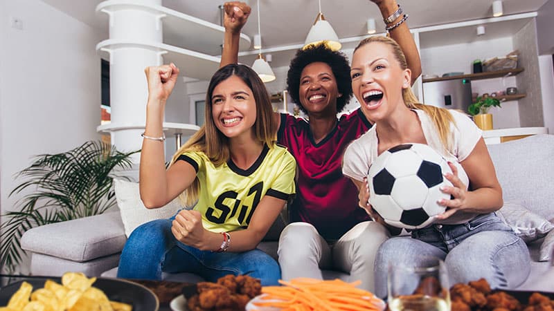 Tres mujeres vestidas con distintas camisetas de sus equipos celebran frente a una mesa con snacks para el partido. Una de ellas sostiene un balón de fútbol.