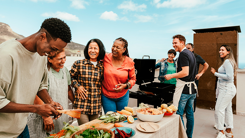 A diverse group of people enjoying an outdoor cookout standing next to a grill and a table with vegetables, fruits, and bread.