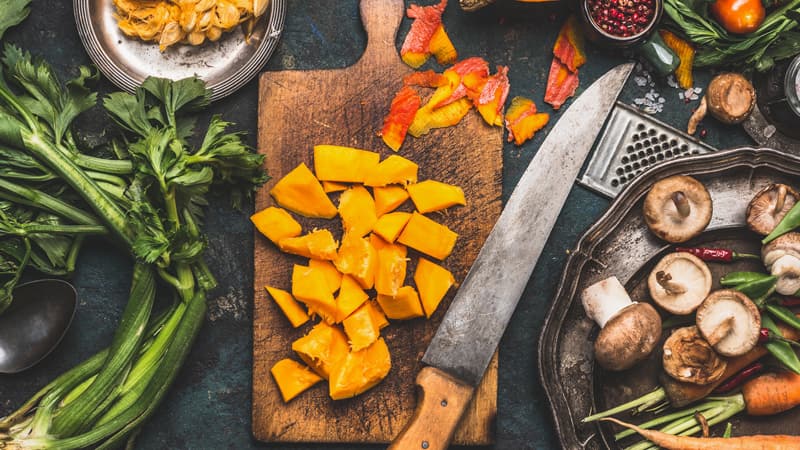 Cutting board with diced pumpkin and knife next to it, surrounded by leafy greens, sliced mushrooms, whole carrots, cherry tomatoes, half cut acorn. Everything is placed on a dark tabletop.