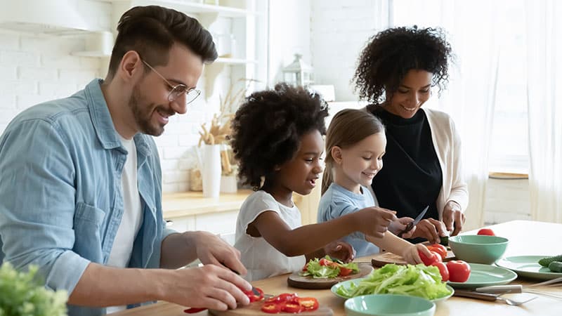 A Mom, dad and two young daughters in a sunlit kitchen, smiling as they prepare a meal together.