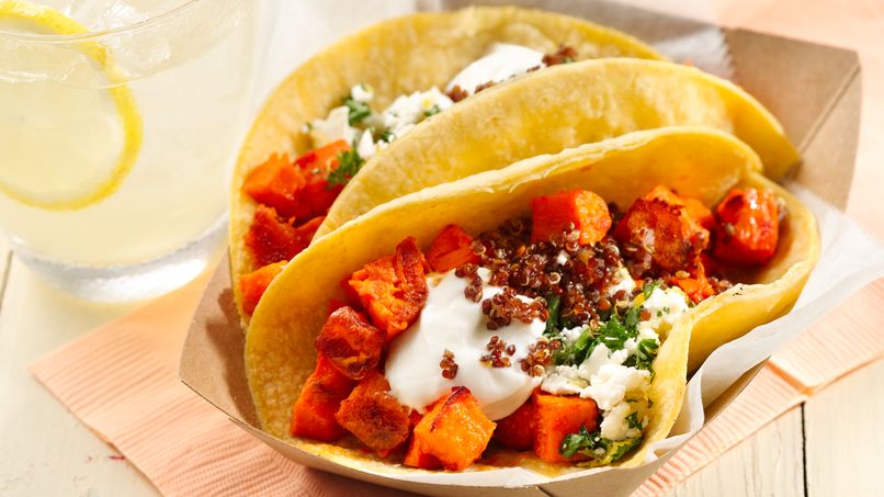 Dos tacos de tortilla de maíz rellenos con camote asado cortado en cubitos, pasta harissa, queso feta desmoronado, perejil picado y espolvoreados con quinoa