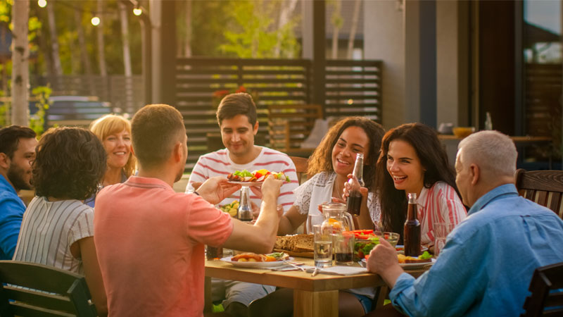 Grupo de hombres y mujeres sentados alrededor de una mesa de jardín, divirtiéndose y disfrutando una comida al aire libre al atardecer.