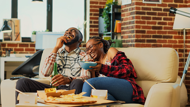 An African American man and woman wearing flannel shirts, sitting on a couch in front of a coffee table with a tray of food, inside a brick dwelling.