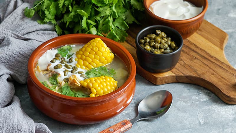 Ajiaco colombiano con trozos de mazorca de maíz servido en plato de barro junto a una cuchara y una tabla con especias y hierbas frescas.”