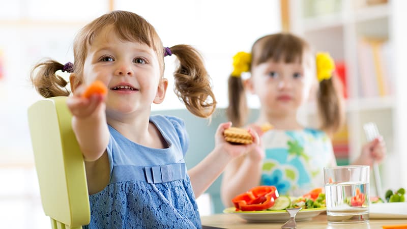 Two little girls with pigtails sitting next to each other at a table with a plate of food and a glass of water in front of them, holding food items in their hands.